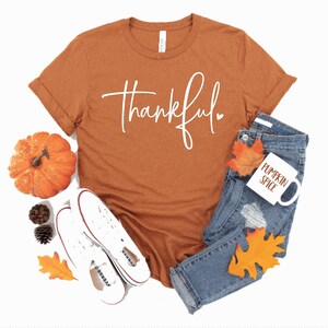 Peut inclure: Un t-shirt couleur rouille avec le mot "thankful" &eacute;crit en blanc en cursive avec un petit c&oelig;ur. Le t-shirt est pos&eacute; sur un fond blanc avec une paire de baskets blanches, une petite citrouille orange et une tasse blanche avec les mots "pumpkin spice" &eacute;crits en orange.