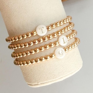 Op de afbeelding: Vier gouden kralenarmbanden met witte parelmoerachtige kralen waarop de letters T, L en G zijn gegraveerd.