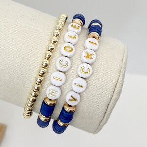 Könnte beinhalten: Drei Perlenarmbänder mit den Namen "Nicole", "Cole" und "Vicki", die in goldenen Buchstaben auf weißen Perlen geschrieben sind. Die Armbänder befinden sich auf einem weißen Ausstellungsständer. Die Armbänder sind blau und goldfarben.