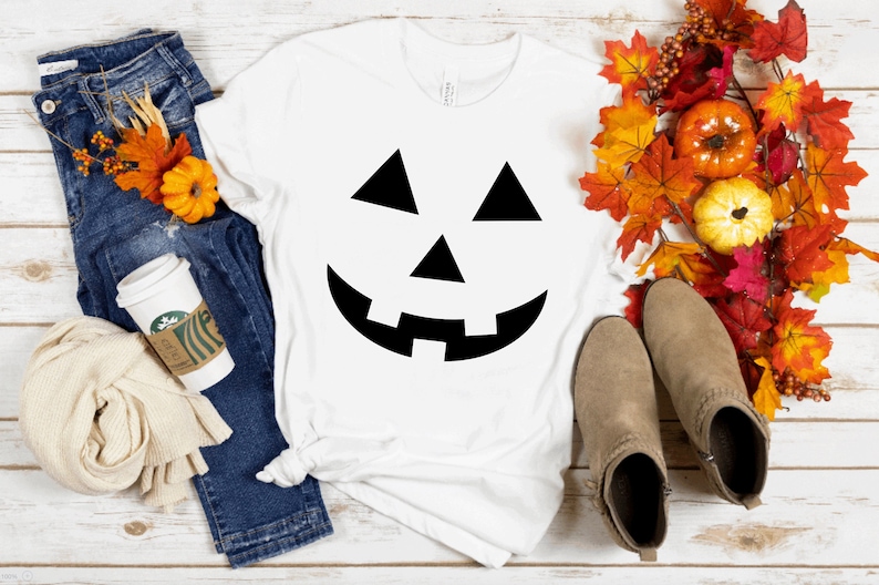 May include: A white short-sleeved t-shirt featuring a black jack-o'-lantern face graphic, paired with blue denim jeans, a beige scarf, a white coffee cup, tan ankle boots, and a decorative arrangement of colorful autumn leaves and pumpkins.