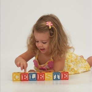 May include: A set of seven square wooden alphabet blocks arranged in a horizontal row to spell the name CELESTE. Each block features a large, bold, sans-serif letter in a contrasting color on a solid background, with smaller letters on the sides.
