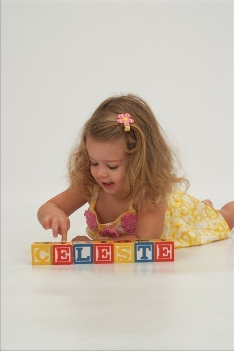 May include: A set of seven square wooden alphabet blocks arranged in a horizontal row to spell the name CELESTE. Each block features a large, bold, sans-serif letter in a contrasting color on a solid background, with smaller letters on the sides.