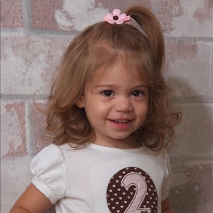 May include: A white short-sleeved shirt featuring a brown circular applique with a white heart pattern and a large light pink number two in the center. A small pink flower hair clip with a brown center is attached to the hair at the top.
