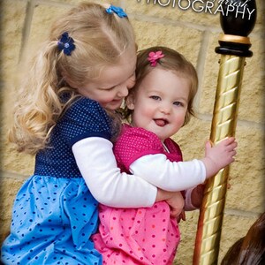 May include: Two children with blonde and brown hair wearing polka dot dresses in blue and pink, each with a small flower hair clip, hugging a gold carousel pole with a black finial. The older child wears a blue dress and the younger a pink dress.
