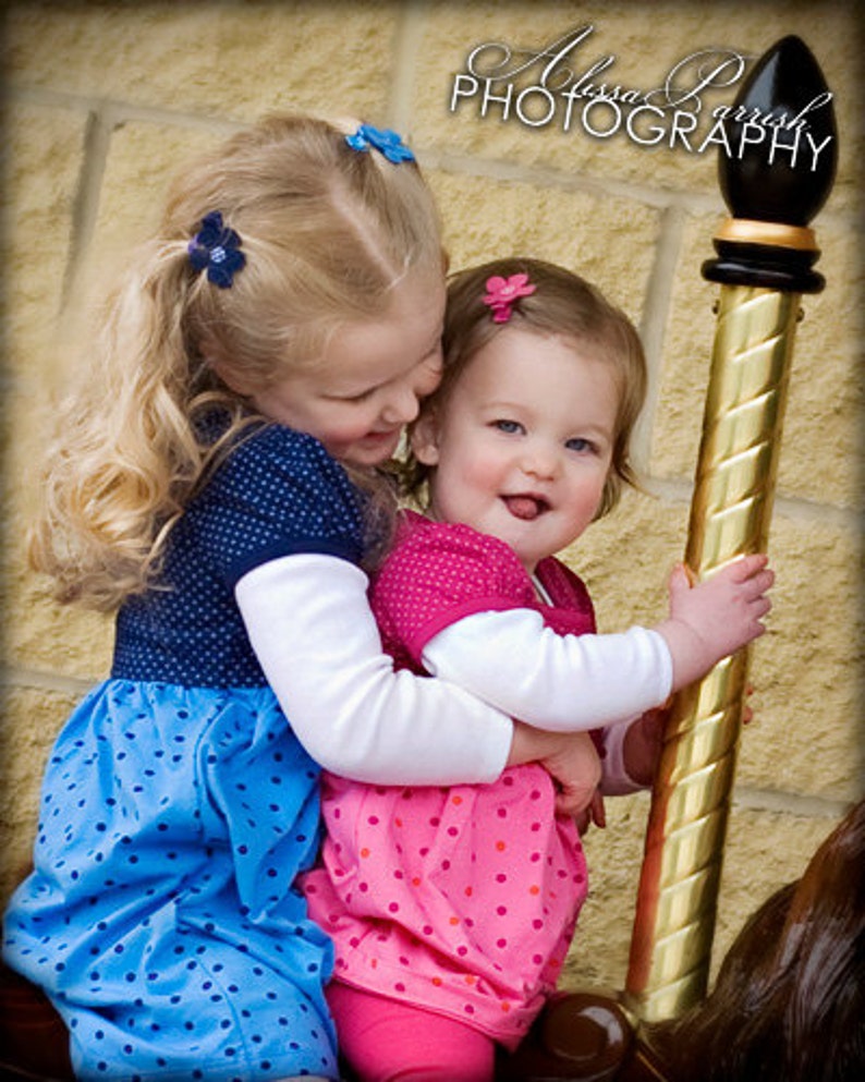 May include: Two children with blonde and brown hair wearing polka dot dresses in blue and pink, each with a small flower hair clip, hugging a gold carousel pole with a black finial. The older child wears a blue dress and the younger a pink dress.
