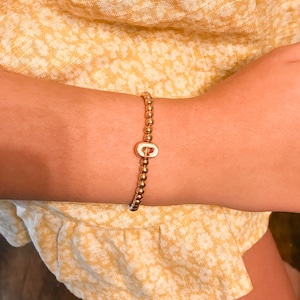 Puede incluir: Un brazalete de cuentas doradas con un dije dorado en forma de la letra "C".