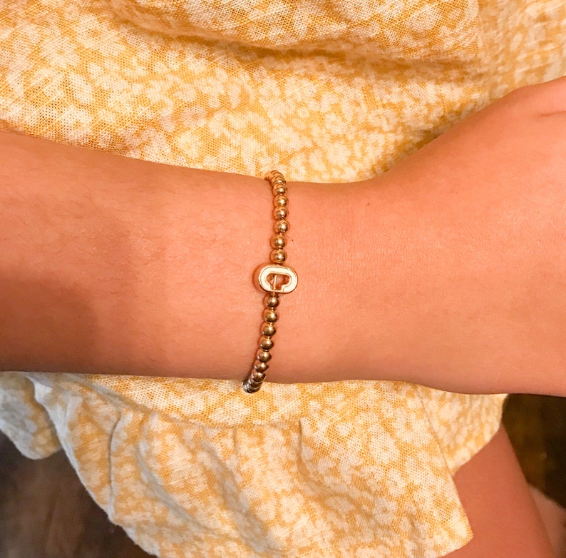 Puede incluir: Un brazalete de cuentas doradas con un dije dorado en forma de la letra "C".