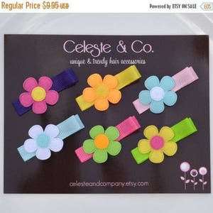 May include: A collection of colorful flower hair clips on a dark background. Each clip features a felt flower in various colors like pink, yellow, blue, and green, attached to a matching ribbon. The brand name "Celeste & Co." is visible.