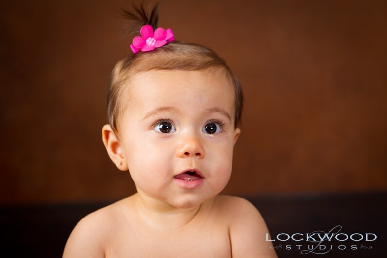 May include: A young child with brown hair styled in a small topknot, adorned with a bright pink flower hair accessory featuring a white checkered center. The child has brown eyes and is looking forward with an open mouth expression.