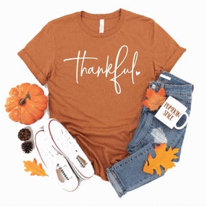 Peut inclure: Un t-shirt couleur rouille avec le mot "thankful" &eacute;crit en blanc en cursive avec un petit c&oelig;ur. Le t-shirt est pos&eacute; sur un fond blanc avec une paire de baskets blanches, une petite citrouille orange et une tasse blanche avec les mots "pumpkin spice" &eacute;crits en orange.