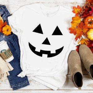 May include: A white short-sleeved t-shirt featuring a black graphic of a jack-o'-lantern face with two triangular eyes, a triangular nose, and a wide, toothy grin. The shirt is displayed alongside blue denim jeans, tan ankle boots, and a scarf.