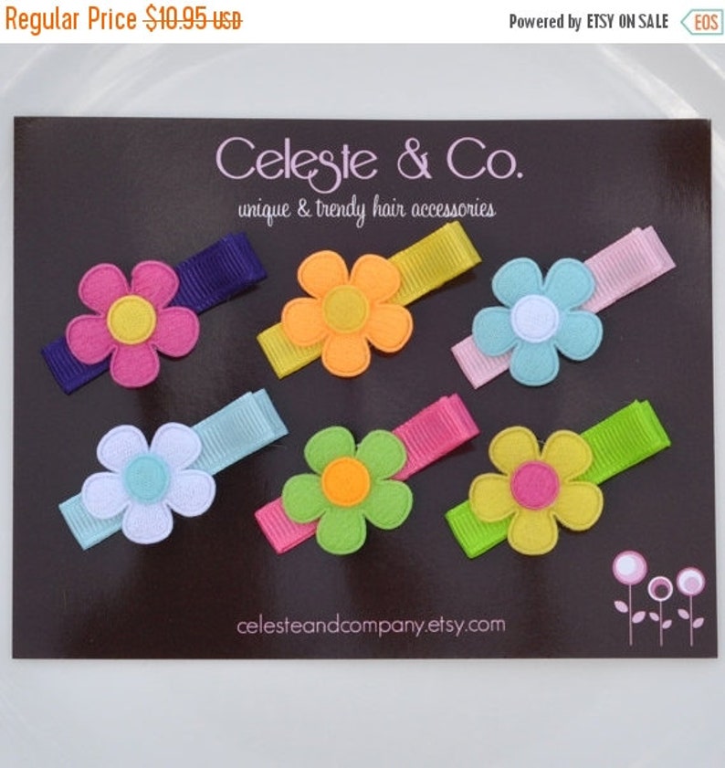 May include: A collection of colorful flower-shaped hair clips. Each clip features a felt flower in shades of pink, orange, blue, green, and yellow, with a contrasting center. The clips are attached to grosgrain ribbon in various colors. The text "Celeste & Co." is visible.
