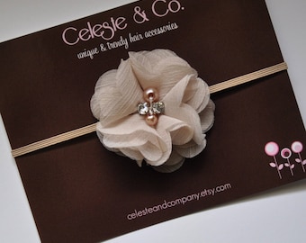 May include: A beige fabric flower with a rhinestone and pearl embellishment is attached to a nude elastic headband. The headband is on a brown background with the text "Celeste & Co. unique & trendy hair accessories" and the website "celesteandcompany.etsy.com".