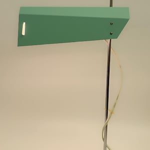 May include: A mint green desk lamp with a rectangular base and a long, angled shade. The lamp has a chrome pole and a visible power cord. The shade has a small, rectangular light opening.