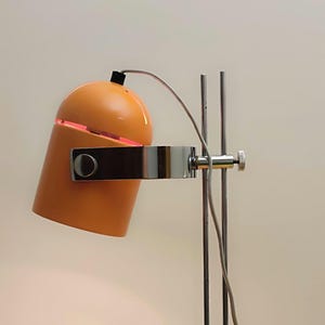 May include: An orange desk lamp with a cylindrical shade and a chrome-colored metal arm and stand. The lamp's shade has a slit that emits a red glow. The lamp is a modern design.
