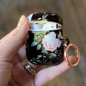 May include: A black and floral patterned Airpod case with a rose gold ring attached. The case is open and the inside is white.