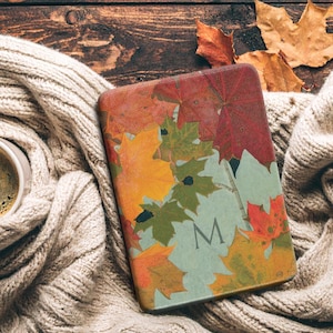 May include: A fall-themed tablet case featuring a colorful leaf design with the letter "M" on a light blue background. The case rests on a cozy, cream-colored knitted scarf, next to a cup of coffee and dried leaves.