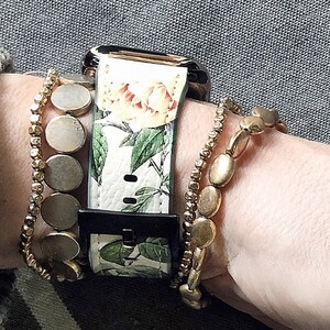 Op de afbeelding: Een witte Apple Watch met een band met bloemenprint. Het horloge wordt gedragen op een pols met gouden kralen armbanden.