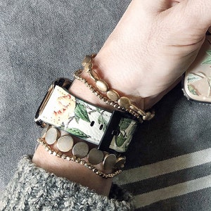 Op de afbeelding: Een wit en groen bloemenpatroon horlogebandje met een zwarte gesp. Het horlogebandje wordt gedragen op een pols met meerdere gouden kralen armbanden.