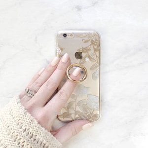 May include: A gold phone case with a floral design and a gold ring holder. The phone case is being held by a hand with a ring on the finger.