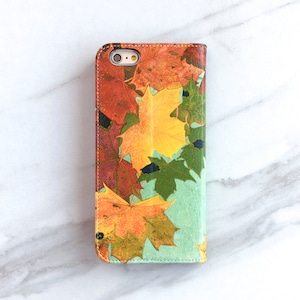 May include: A phone case with a colorful autumn leaf design. The case is made of leather and has a stitched edge. The leaves are in shades of red, orange, yellow, and green.