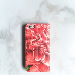 May include: A red and white floral print phone case. The case is a folio style, with a cover that folds over the screen. The floral design features large, detailed peony blooms. The case is designed to fit a smartphone.