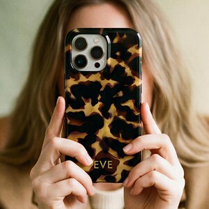 May include: A smartphone case featuring a glossy tortoiseshell pattern in shades of brown, black, and amber. The case has a cutout for a triple-lens camera system and the name EVE is printed in gold lettering near the bottom center of the back.