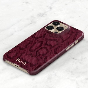 Peut inclure: Une coque de téléphone en peau de serpent bordeaux avec un objectif d'appareil photo doré et le nom "Beth" en relief sur le bas.