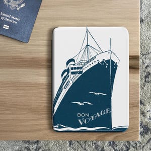 May include: A rectangular white case featuring a dark blue graphic illustration of a large cruise ship sailing forward. The text BON VOYAGE is printed in dark blue below the ship, accompanied by two small birds flying near the bow of the vessel.