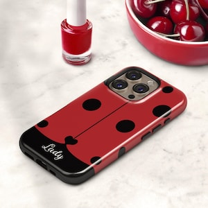 May include: A red and black ladybug-themed phone case with black polka dots and the word "Lady" in cursive. The case has a glossy finish and is designed to fit a smartphone. A red nail polish bottle and a bowl of cherries are in the background.