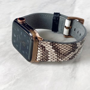May include: A rose gold Apple Watch with a snakeskin patterned band in shades of white, brown, and black. The watch face displays the time. The band has a gray leather backing and a gold buckle.