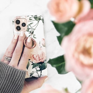 May include: A white phone case with a floral design featuring pink roses and green leaves. A gold ring is attached to the case.