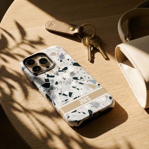 May include: A smartphone case featuring a white, gray, and dark green terrazzo pattern with a horizontal beige stripe across the lower section. The stripe contains the white capital letters JMS. The case has a black camera lens cutout area.