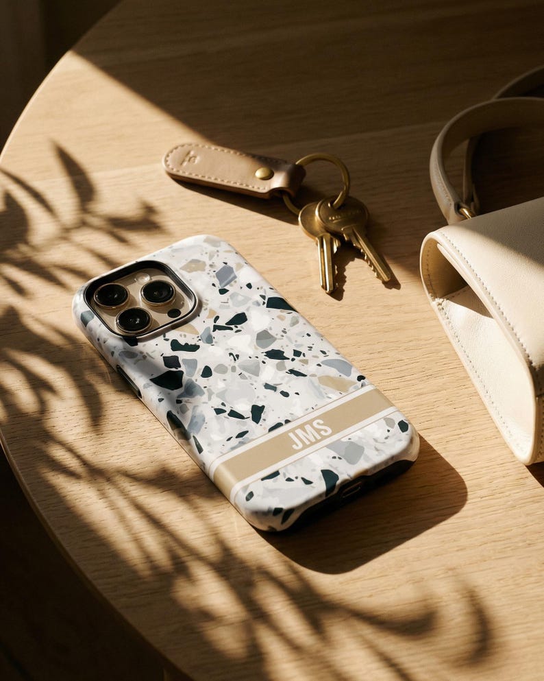 May include: A smartphone case featuring a white, gray, and dark green terrazzo pattern with a horizontal beige stripe across the lower section. The stripe contains the white capital letters JMS. The case has a black camera lens cutout area.