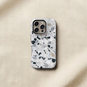 May include: A protective phone case featuring a white, gray, and black terrazzo pattern with scattered geometric shapes. The case has a rectangular cutout at the top for a triple-lens camera system, showing three circular lenses and a flash.