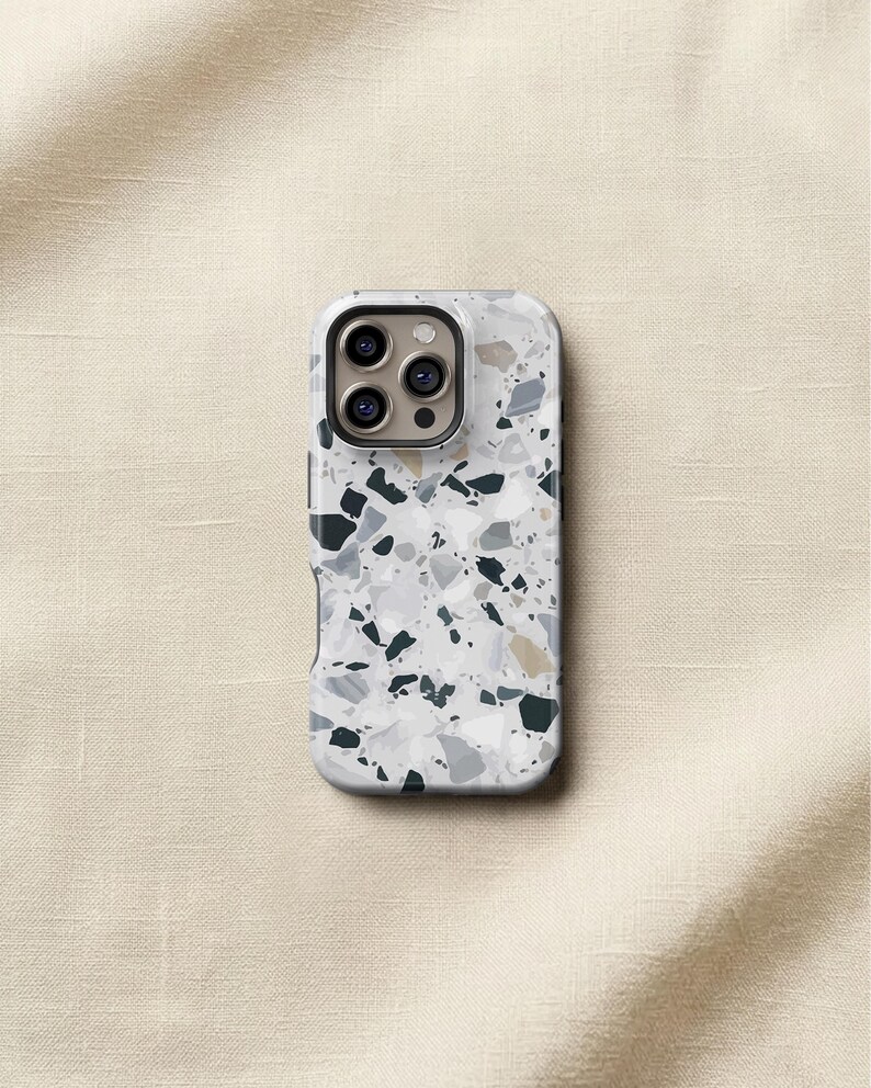 May include: A protective phone case featuring a white, gray, and black terrazzo pattern with scattered geometric shapes. The case has a rectangular cutout at the top for a triple-lens camera system, showing three circular lenses and a flash.