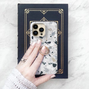May include: A white phone case with a terrazzo pattern and a gold ring holder. The case is on a dark blue book with a gold border.