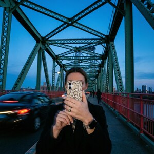 May include: A smartphone featuring a beige and brown snakeskin patterned protective case, held up to capture a photograph on a green metal bridge with red railings at dusk, with a blurred car passing by on the left side of the frame.