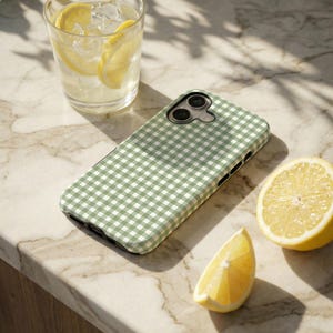 May include: A green and white gingham patterned phone case rests on a marble surface, next to a glass of lemonade and a halved lemon. The case has a camera cutout and a sleek design, suggesting a focus on style and protection.