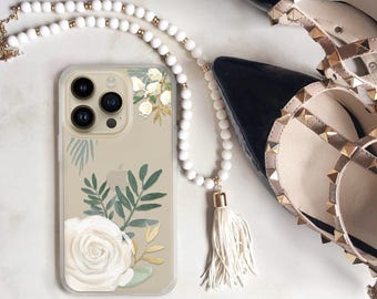 Puede incluir: Una funda transparente para smartphone con una rosa blanca, hojas verdes y detalles dorados. Junto a ella hay un collar largo de cuentas blancas con una borla y un par de zapatos de tacón negros con tachuelas doradas.