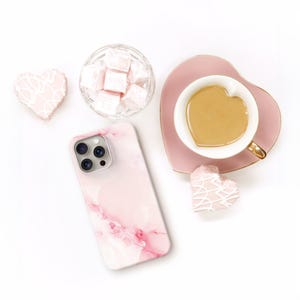 May include: A pink marble-patterned smartphone case, a heart-shaped ceramic cup filled with coffee on a matching saucer, a glass bowl of cubed pink treats, and two heart-shaped cookies with white icing drizzle arranged on a white surface.