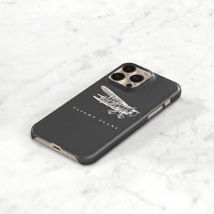 Peut inclure: Coque d'iPhone gris fonc&eacute; avec une illustration d'avion argent&eacute;e et les mots "FLIGHT PLANS" en blanc. L'&eacute;tui a un cadre dor&eacute; et une d&eacute;coupe pour cam&eacute;ra &agrave; trois objectifs.