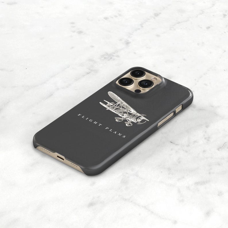 Peut inclure: Coque d'iPhone gris fonc&eacute; avec une illustration d'avion argent&eacute;e et les mots "FLIGHT PLANS" en blanc. L'&eacute;tui a un cadre dor&eacute; et une d&eacute;coupe pour cam&eacute;ra &agrave; trois objectifs.
