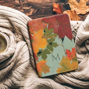 May include: A rectangular e-reader case featuring a vibrant autumn leaf pattern in shades of red, orange, yellow, and green. The design includes a prominent capital letter M centered near the bottom, set against a light green background.