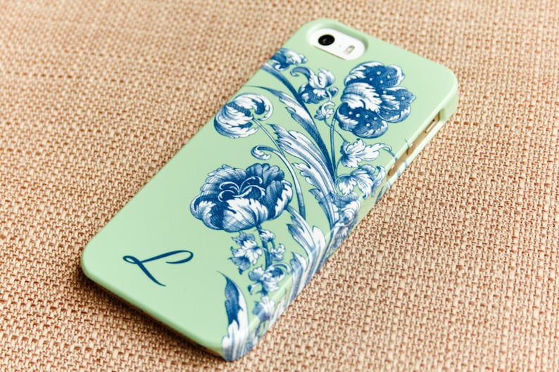May include: A mint green iPhone case with a blue and white floral design and the letter "L" printed on the back.