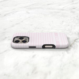 May include: A pink and white gingham patterned phone case with the name "ASHLEY" printed vertically on the side. The case has a black camera cutout and a black button cutout.