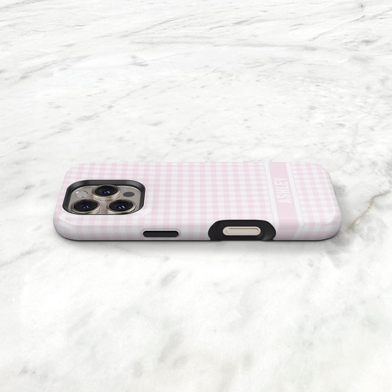 May include: A pink and white gingham patterned phone case with the name "ASHLEY" printed vertically on the side. The case has a black camera cutout and a black button cutout.