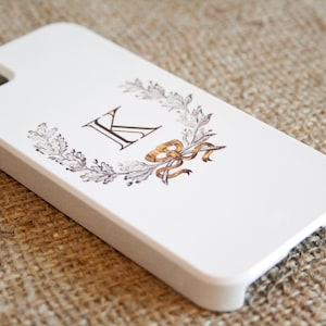 May include: White iPhone case with a black and gold design. The design features a wreath of leaves with a gold ribbon and the letter "K" in the center.