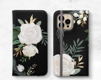 Puede incluir: Funda de teléfono negra con un patrón floral blanco. La funda tiene una rosa blanca, hojas verdes y detalles dorados. La funda tiene un recorte para la cámara y una banda elástica gris.