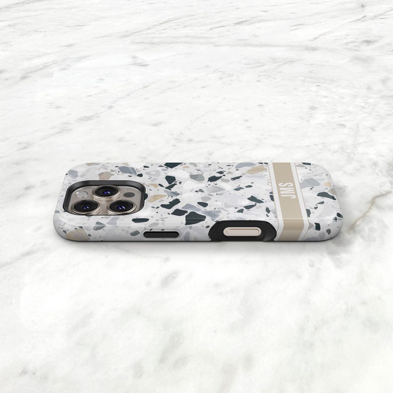 May include: A smartphone case featuring a white, gray, and black terrazzo pattern with a vertical beige stripe on the right side containing the letters JMS in white. The case has a black camera cutout area with three circular lens openings.